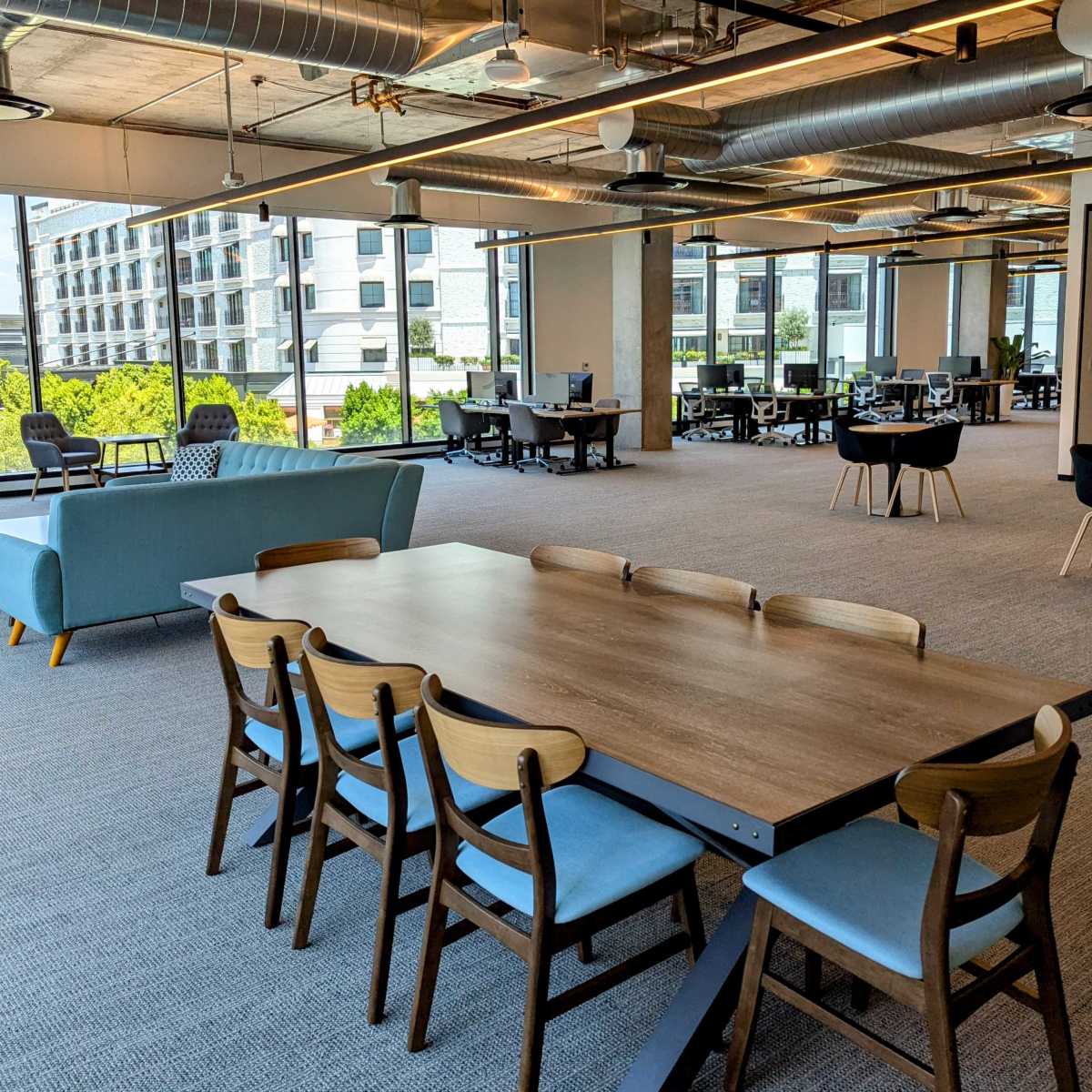 A spacious, modern office interior with a large wooden table surrounded by six chairs, and various workstations in the background.