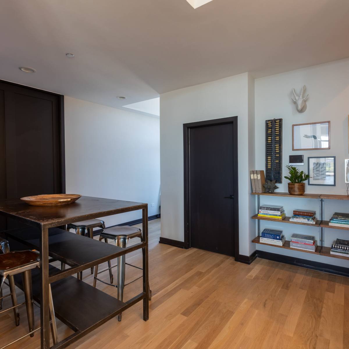 A modern interior space featuring a wooden table with stools, an exit sign, and a wall shelf filled with books.