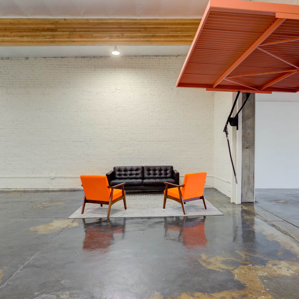 A black sofa and two orange chairs are positioned on a rug in an open, minimalistic space with concrete flooring and a white brick wall.