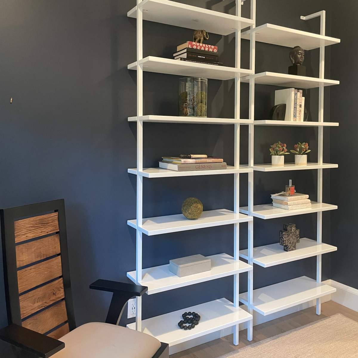A minimalist white shelving unit against a dark blue wall, with various books and decorative objects displayed on the shelves, accompanied by a wooden chair in the foreground.