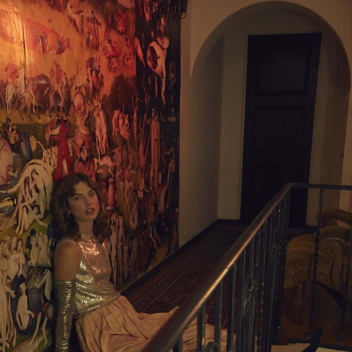 A woman in a sequined top and long skirt sits against a wall adorned with an elaborate mural, in a softly lit hallway.
