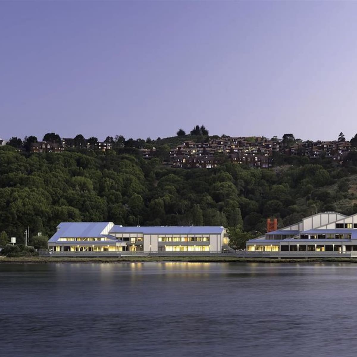 A modern building complex alongside a calm body of water, with a hillside covered in greenery and houses in the background.