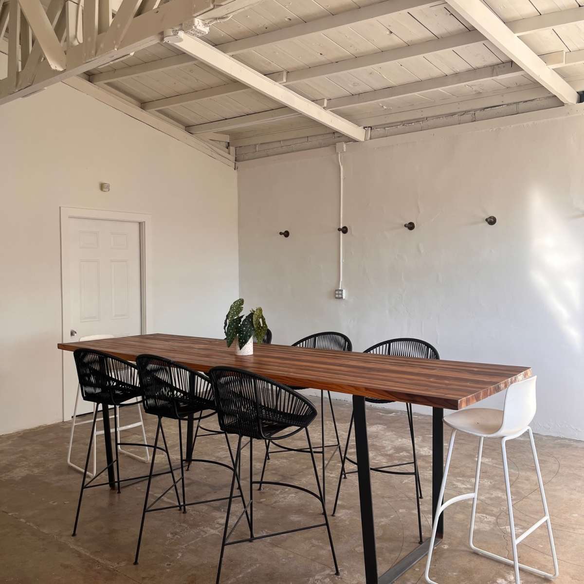 A long wooden table with black and white high chairs is set in a minimalistic room with a white ceiling and walls.