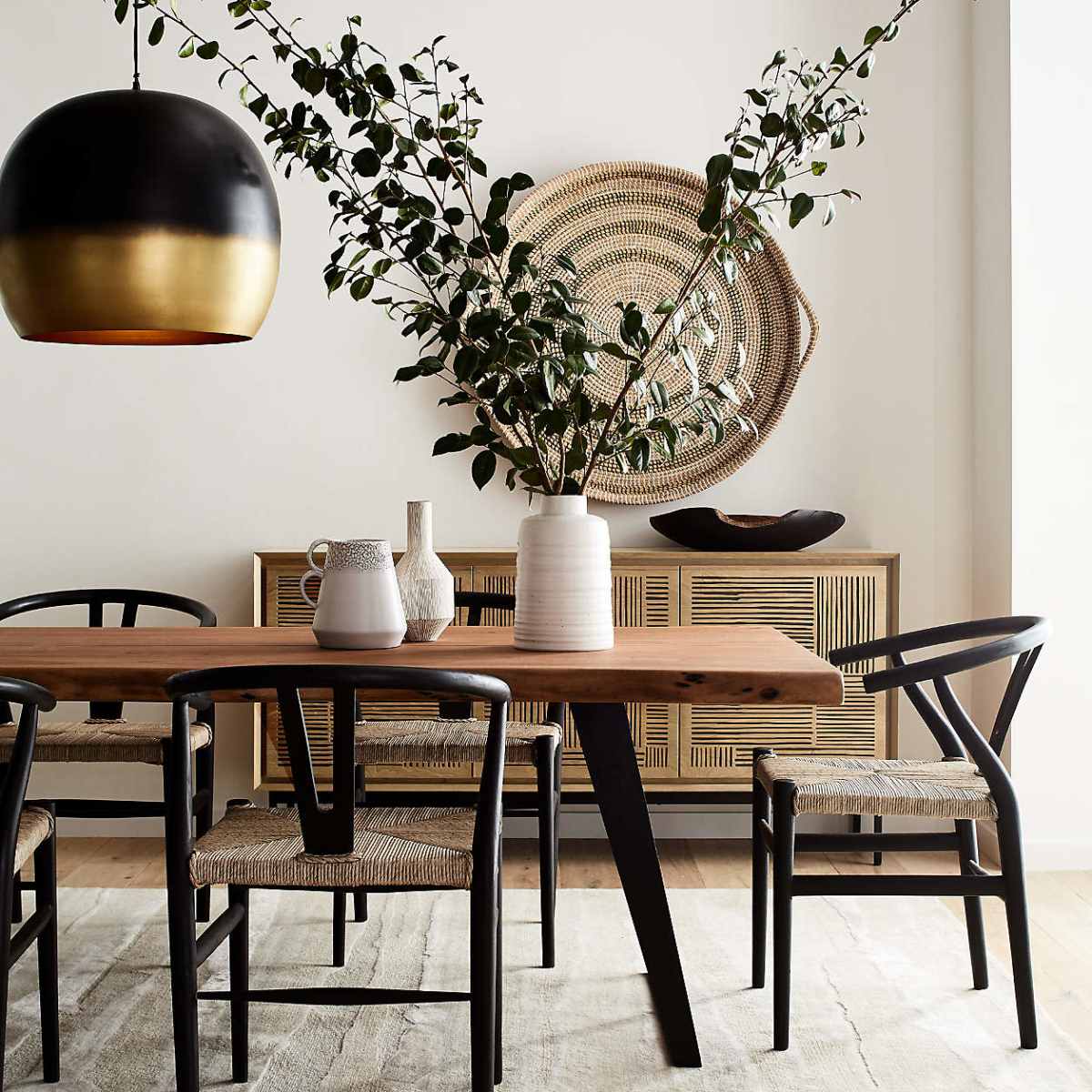 A wooden dining table with black chairs is set against a light wall, featuring a large pendant light above and a vase with branches as a centerpiece.