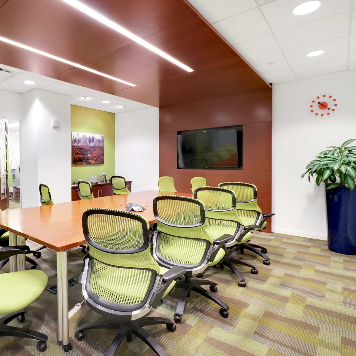 A modern conference room with a large wooden table surrounded by green ergonomic chairs, featuring a wall-mounted television and a large indoor plant.