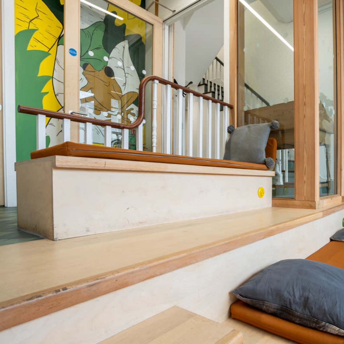 A bright indoor space with wooden steps, cushions, and a glass partition featuring a green mural of a cartoon animal.