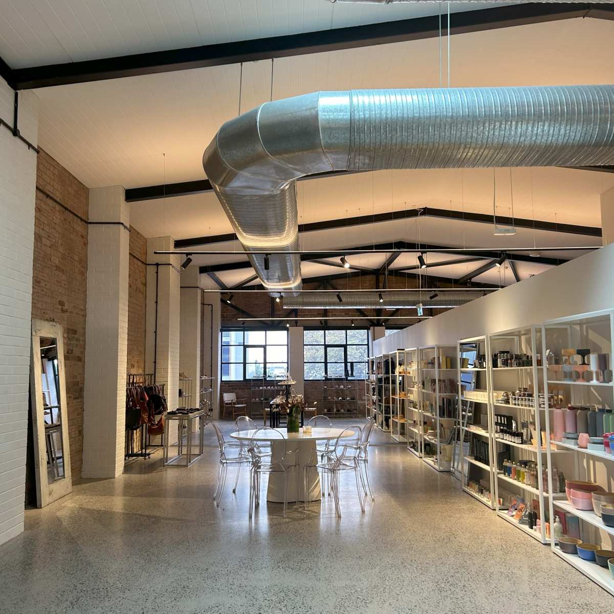 A spacious interior featuring a large table surrounded by chairs, with shelves displaying various products along the walls and industrial-style ductwork overhead.