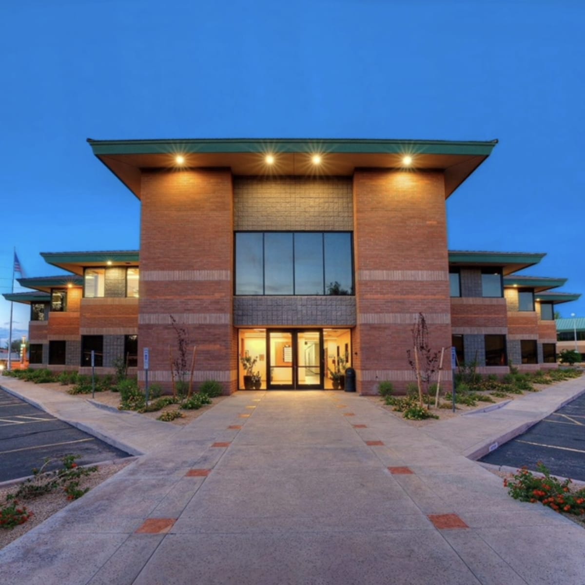 A modern brick building with a flat roof and large windows, flanked by parking spaces and landscaped areas in the front.
