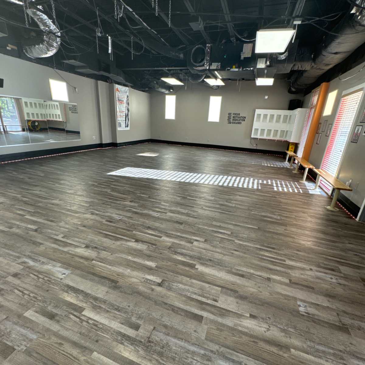 A spacious, empty room with a wooden floor, large windows, and a mirrored wall.