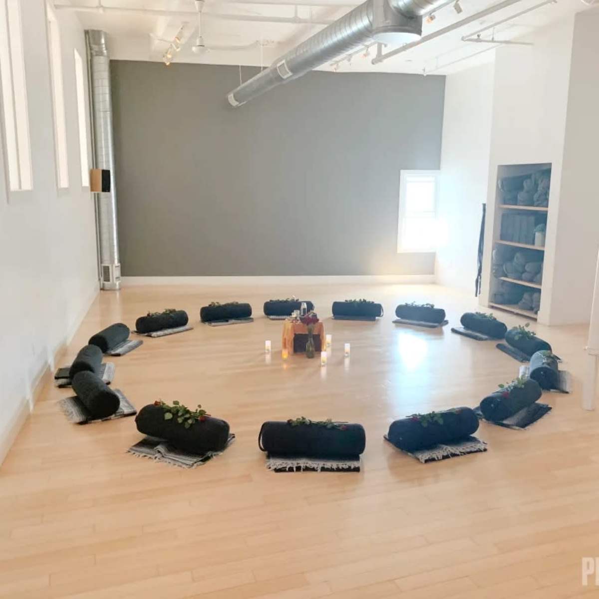 A circular arrangement of black meditation cushions is set up on a wooden floor in a bright, spacious room.