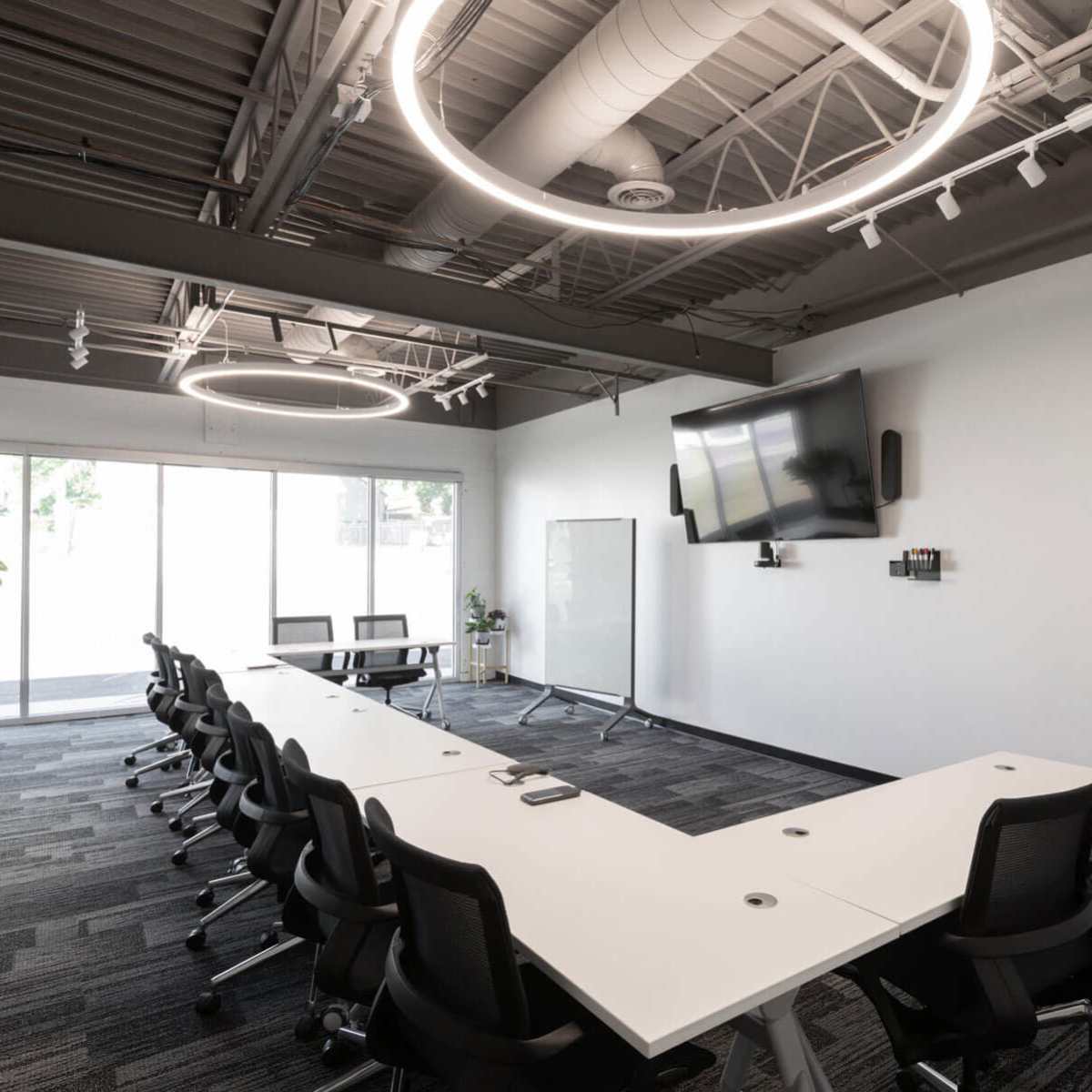 A modern conference room with a long, white table surrounded by black chairs, large windows, and circular light fixtures overhead.