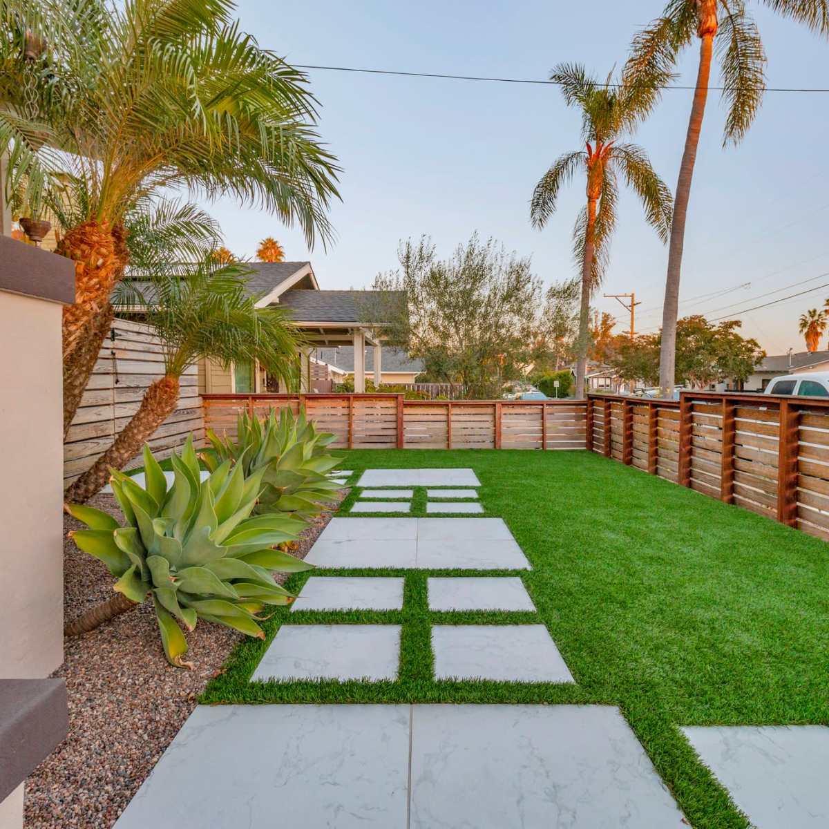 A landscaped backyard with stone pathways, artificial grass, and palm trees, enclosed by wooden fencing.
