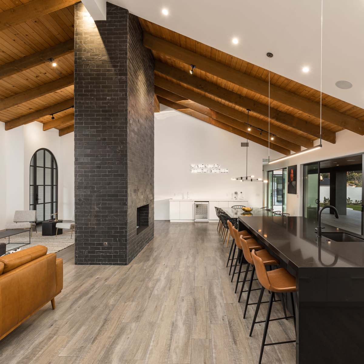 A modern kitchen and living area with a high wooden ceiling, a central black island with bar stools, and a large fireplace set against a black brick wall.