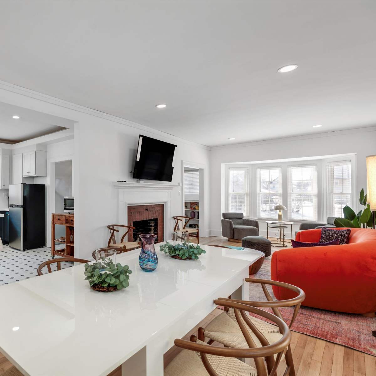 A modern kitchen and dining area features a white table, bar stools, and a living room with a large window and colorful furniture.