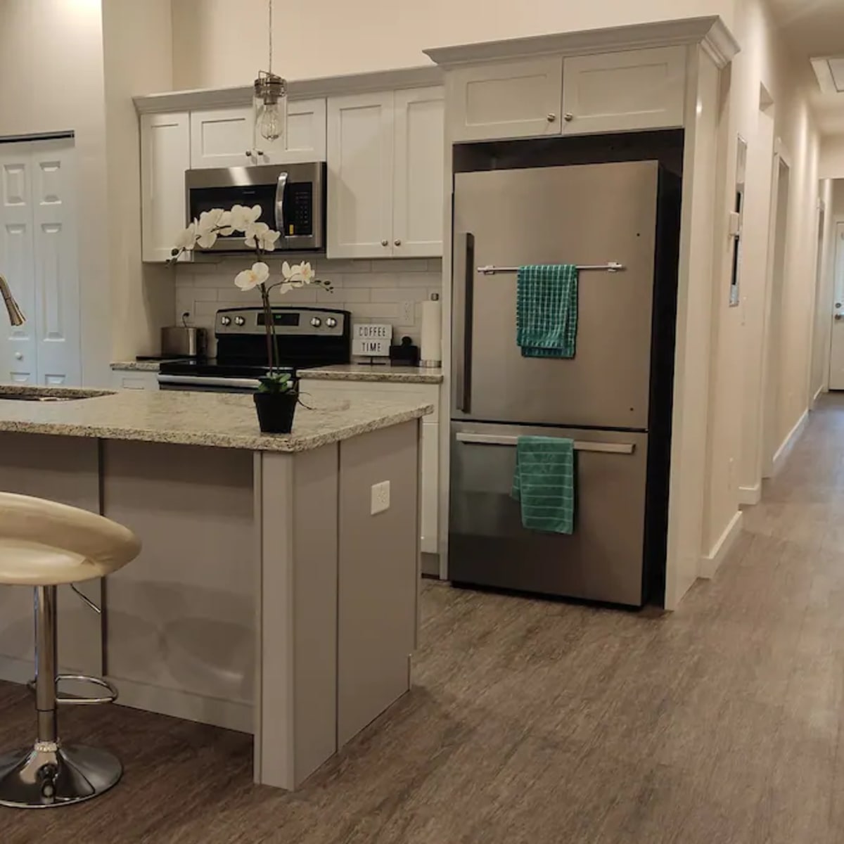 A modern kitchen with bar stools, granite countertops, stainless steel appliances, and a hallway leading to a door at the end.