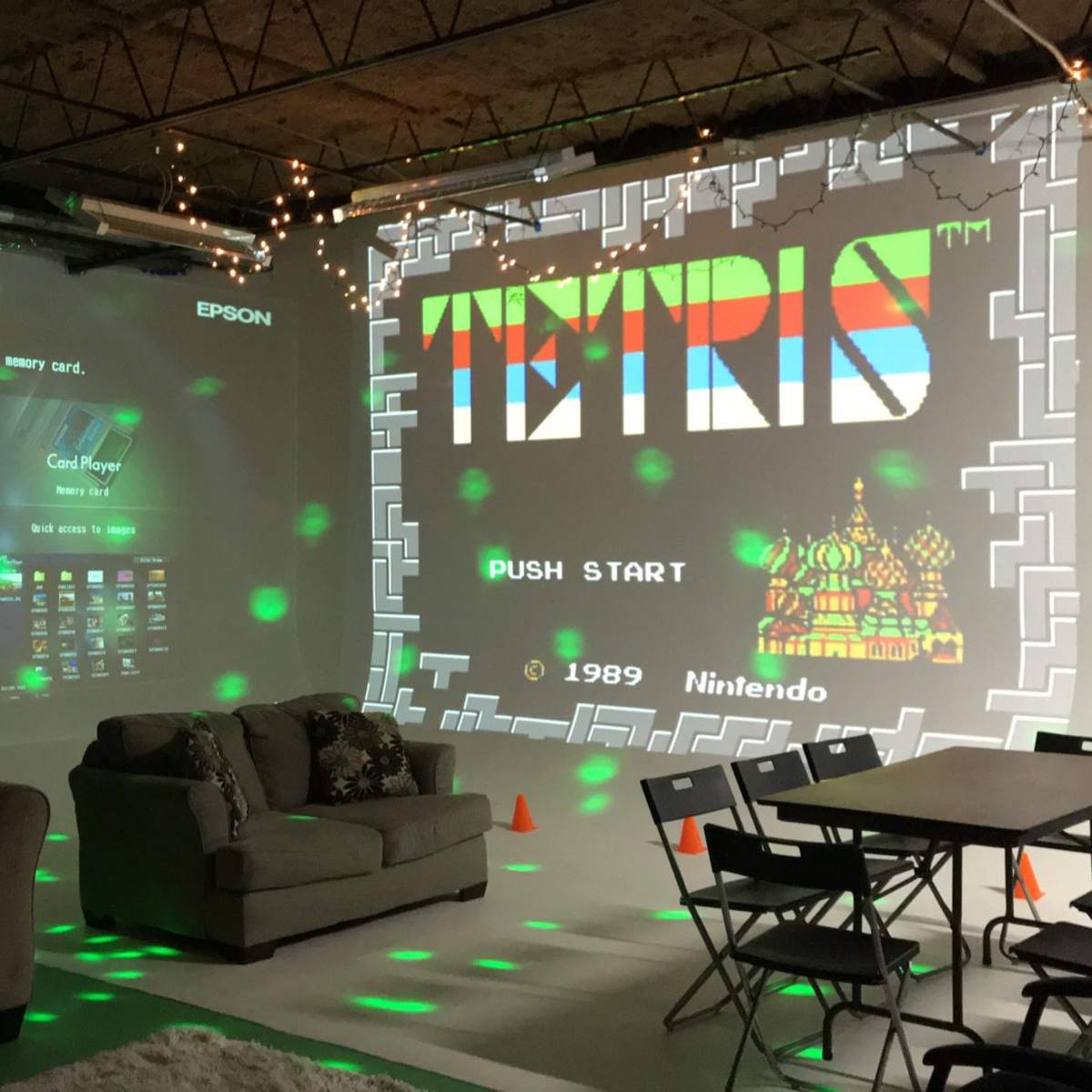 A recreation room with a projector displaying a Tetris game on a wall, alongside a couch and a table set for guests.