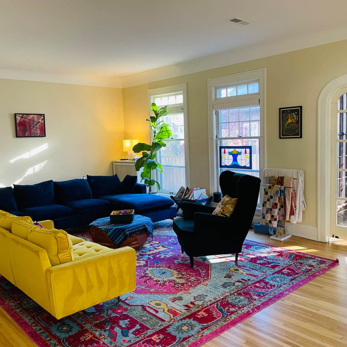 A bright living room features a blue sectional sofa, a yellow couch, and a colorful area rug, with large windows allowing natural light to fill the space.