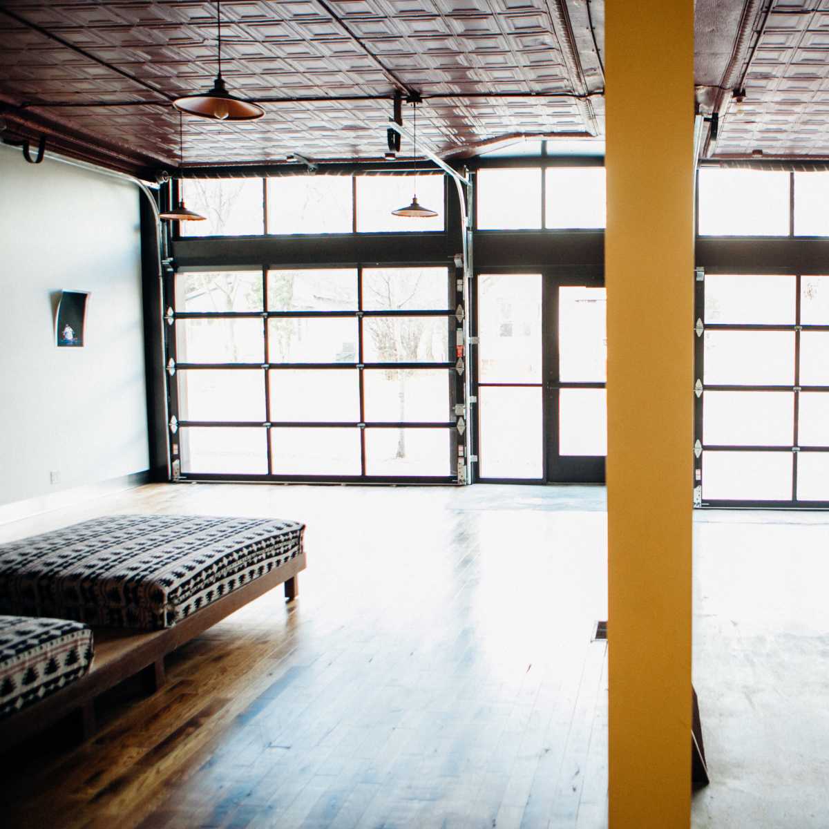 An empty interior space with polished wooden floors, a combination of large glass doors and windows, and two seating areas divided by a yellow column.