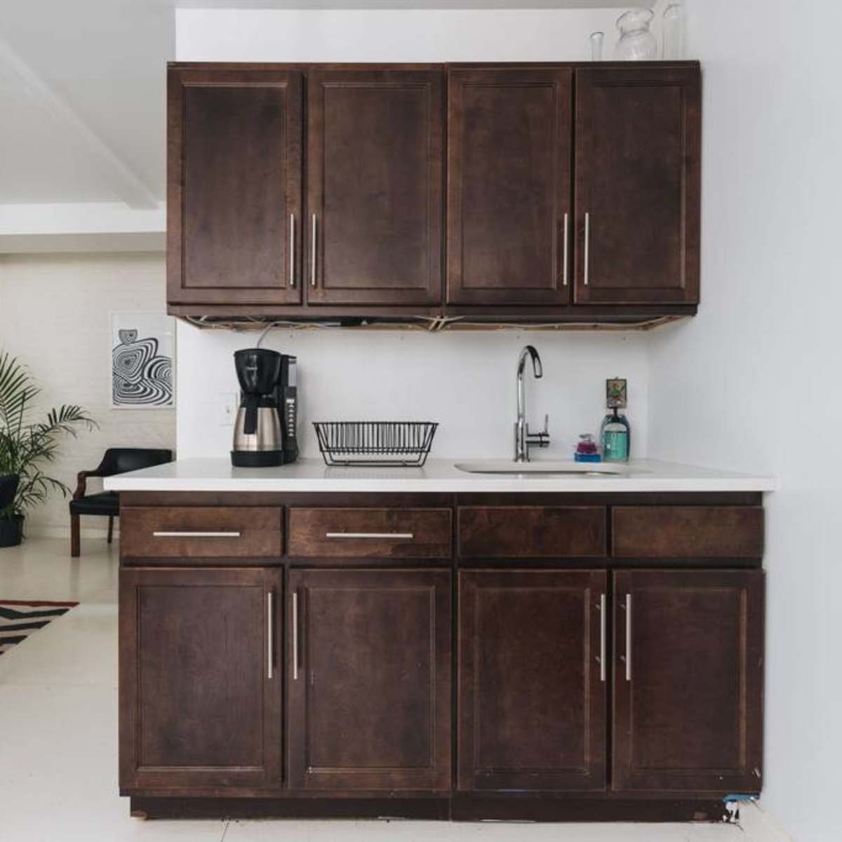 A kitchen area featuring dark wood cabinets, a coffee maker, a sink, and a dining area in the background with a patterned rug.