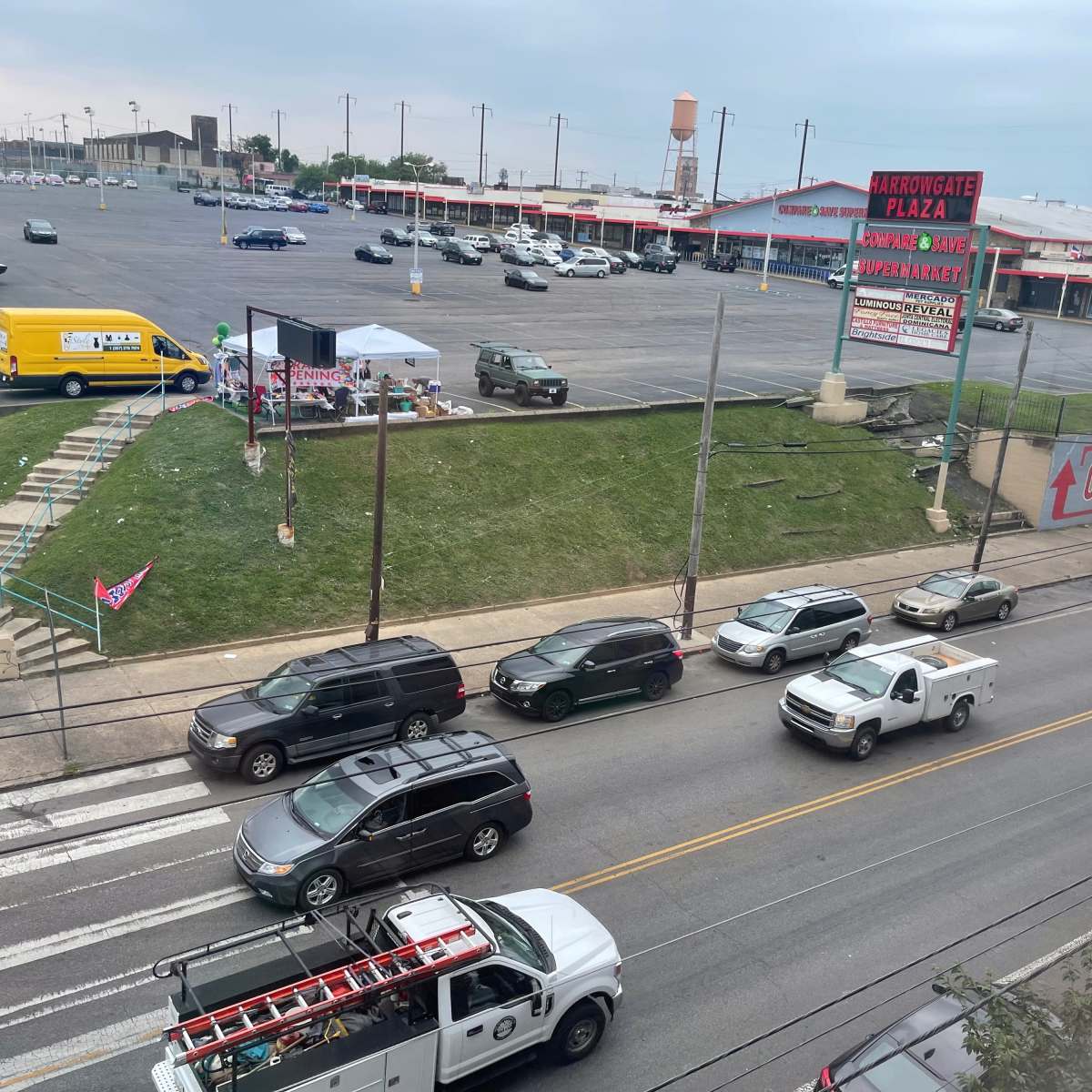 A busy street scene with multiple vehicles passing by a large parking lot and a nearby building displaying signage.