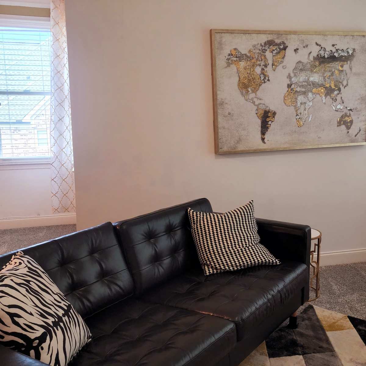 A black leather sofa with a patterned pillow faces a wall featuring a gold-accented world map, under soft lighting from nearby windows.