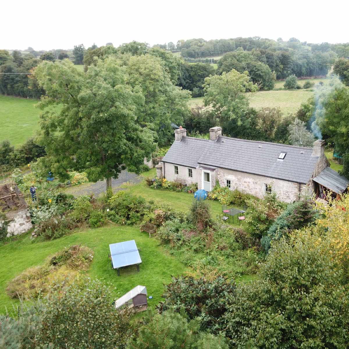 An aerial view shows a stone cottage surrounded by lush greenery and trees, with smoke rising from its chimney.