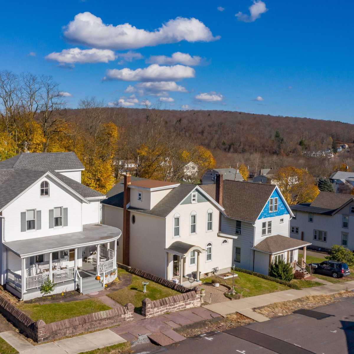 A row of residential houses with varied architectural styles and colors, set against a backdrop of trees and hills under a clear blue sky.