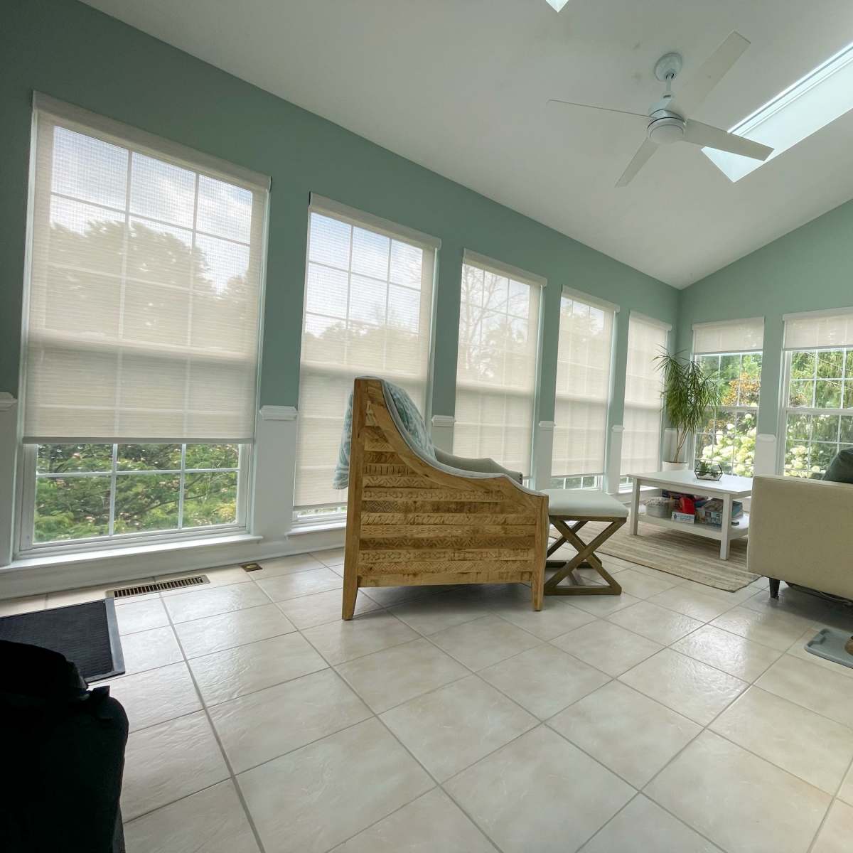 A sunlit sunroom with a wicker chair, a beige sofa, and large windows that allow views of greenery outside.