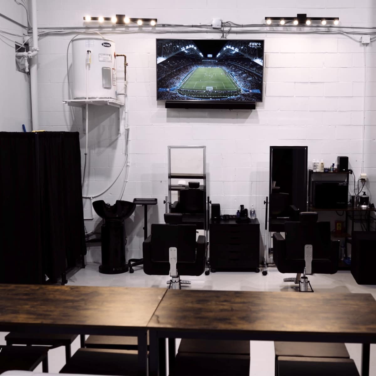A modern salon interior with a large television displaying a sports event, black styling chairs, and a wooden table in the foreground.