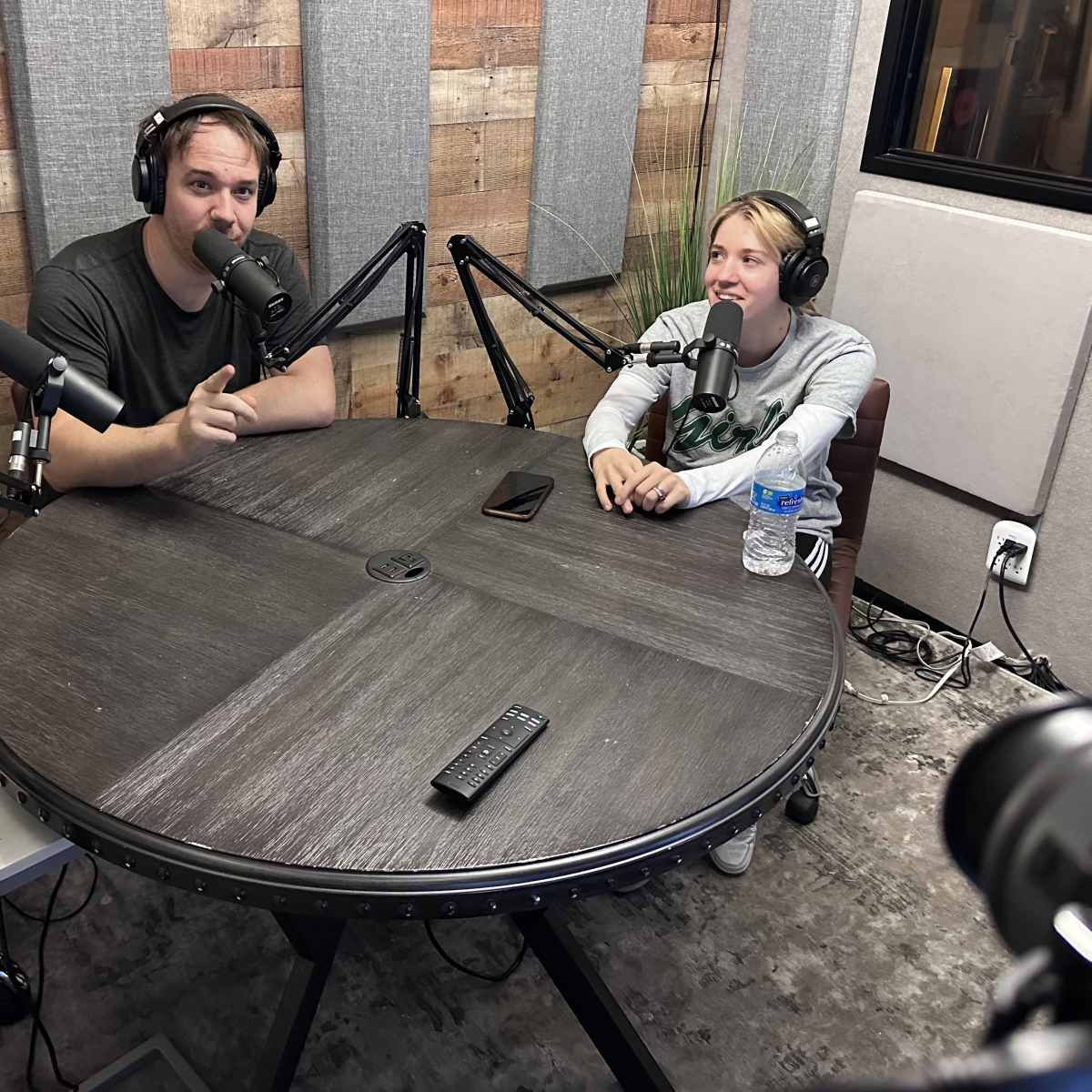 A man and a woman sit at a large round table in a recording studio, each speaking into a microphone while headphones are worn.