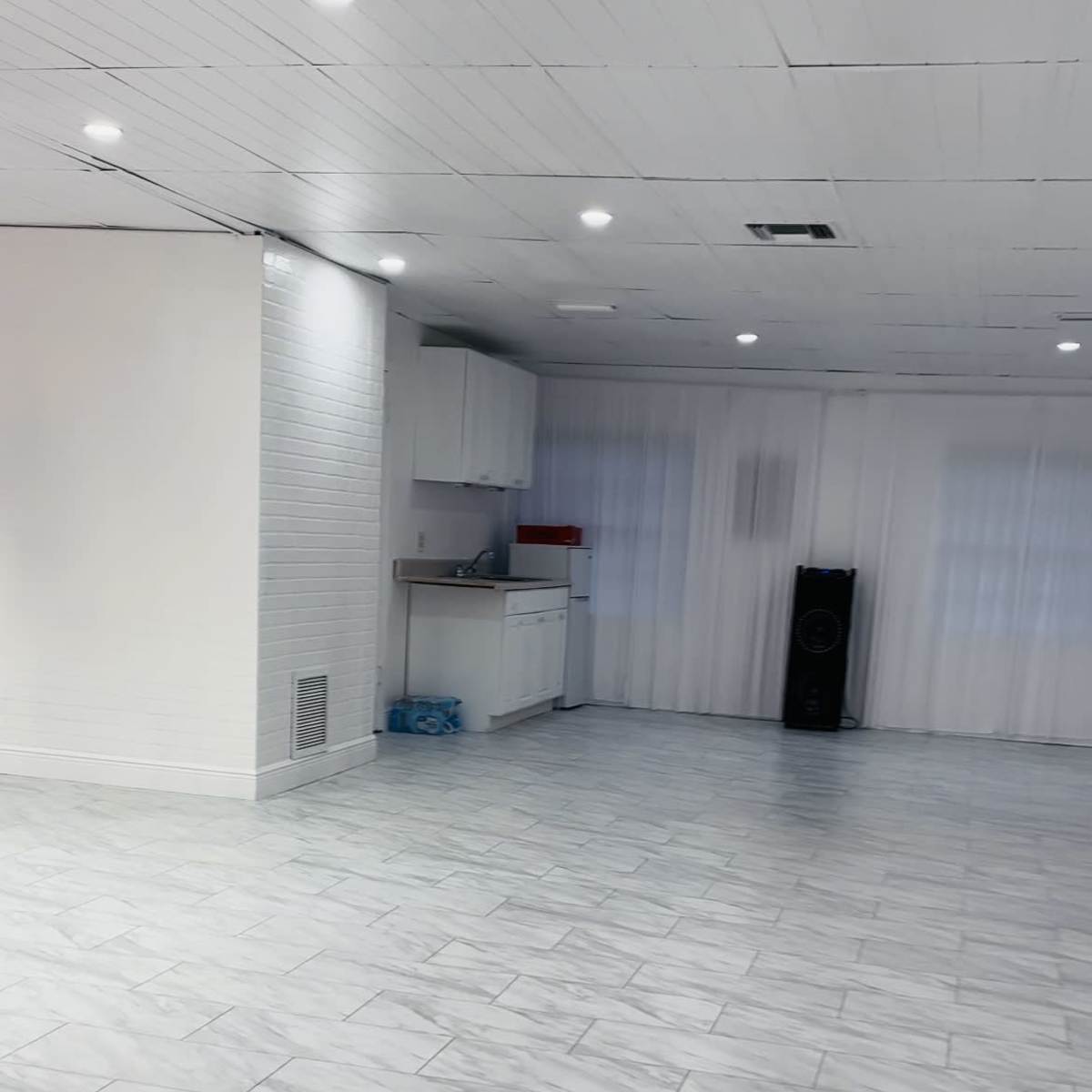 A large, empty room with white tiled flooring, a kitchenette in the corner, and large windows covered with sheer curtains.