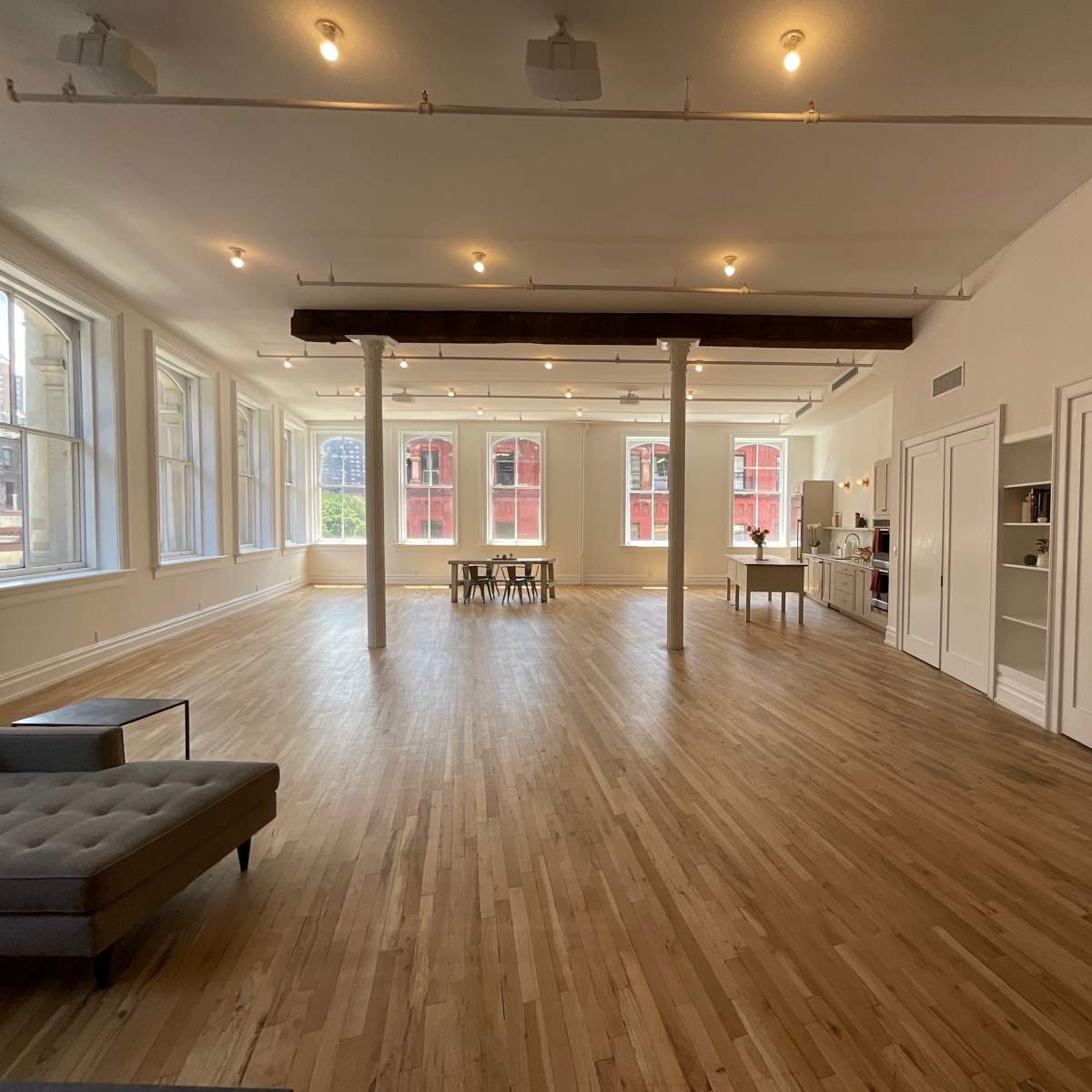 A spacious, well-lit room with wooden floors, large windows, a dining table, and a kitchenette in the corner.