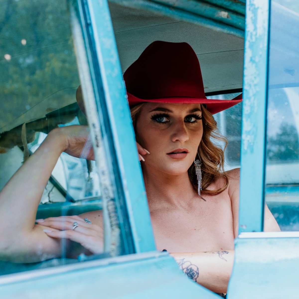 A woman with long hair and a red hat poses inside an old blue vehicle, resting her chin on her hand.