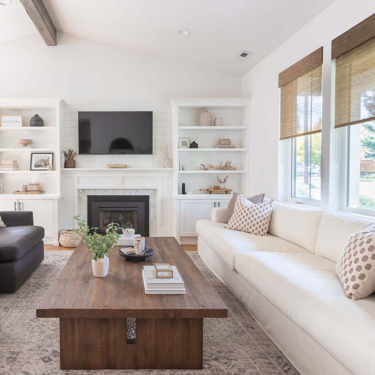 A modern living room features a white couch, dark armchairs, a wooden coffee table, built-in shelving, and a television above a fireplace.