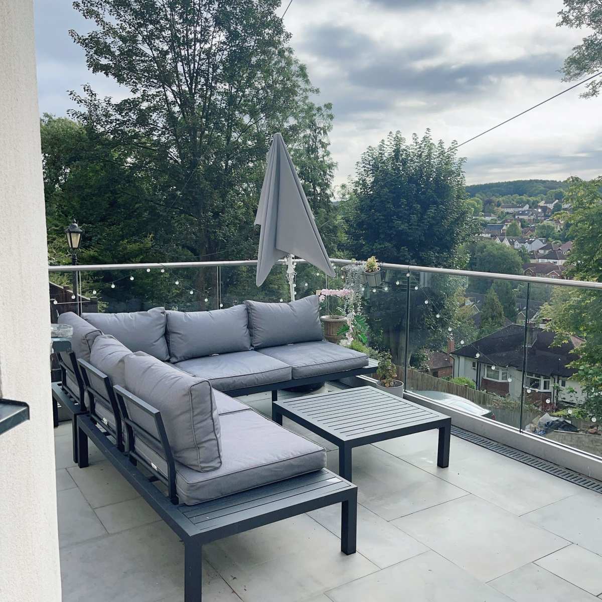 A modern outdoor seating area features gray cushions on black furniture, with a table and an umbrella, overlooking a green landscape.