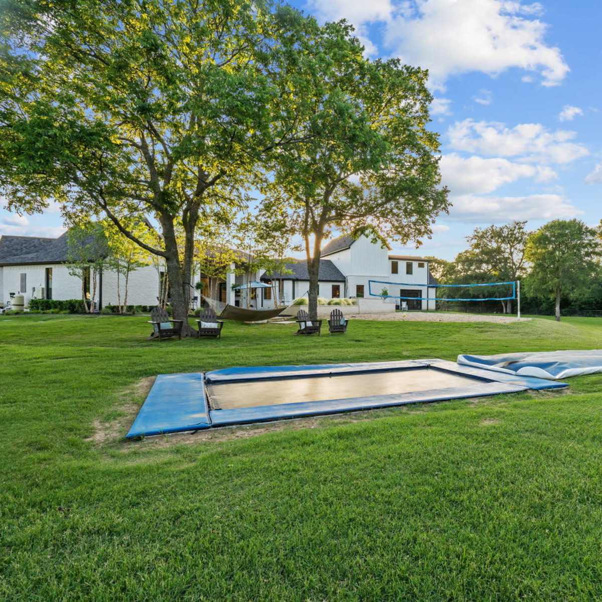 A grassy yard featuring a trampoline and a volleyball net beside a white house and trees in the background.