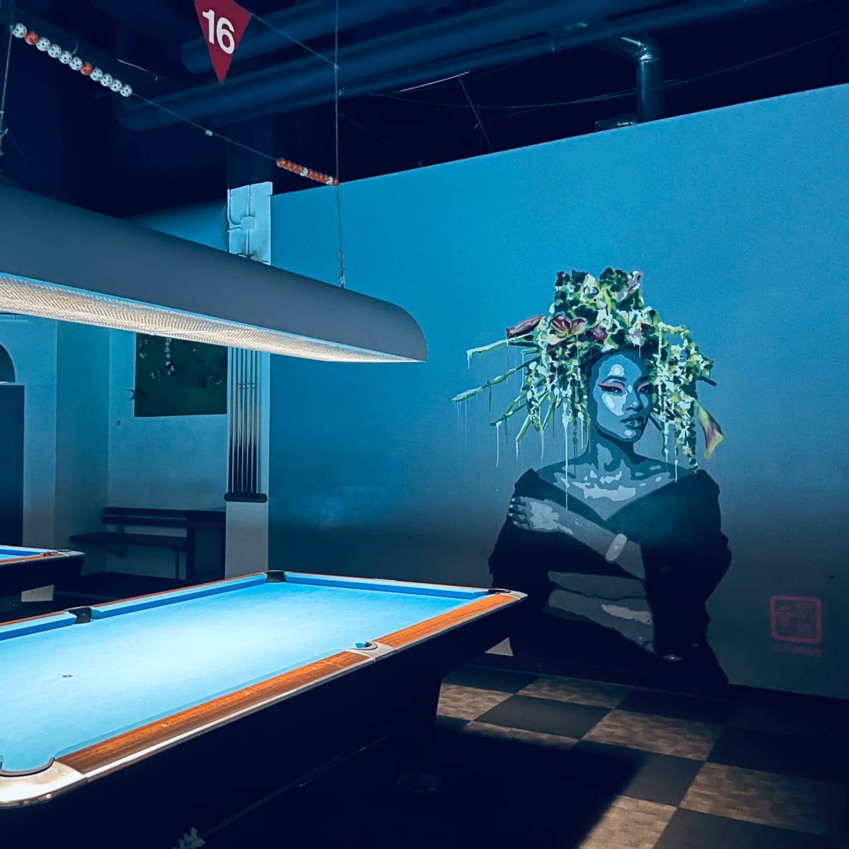 A pool hall featuring a vibrant mural of a woman with greenery on her head next to a blue-felted pool table.