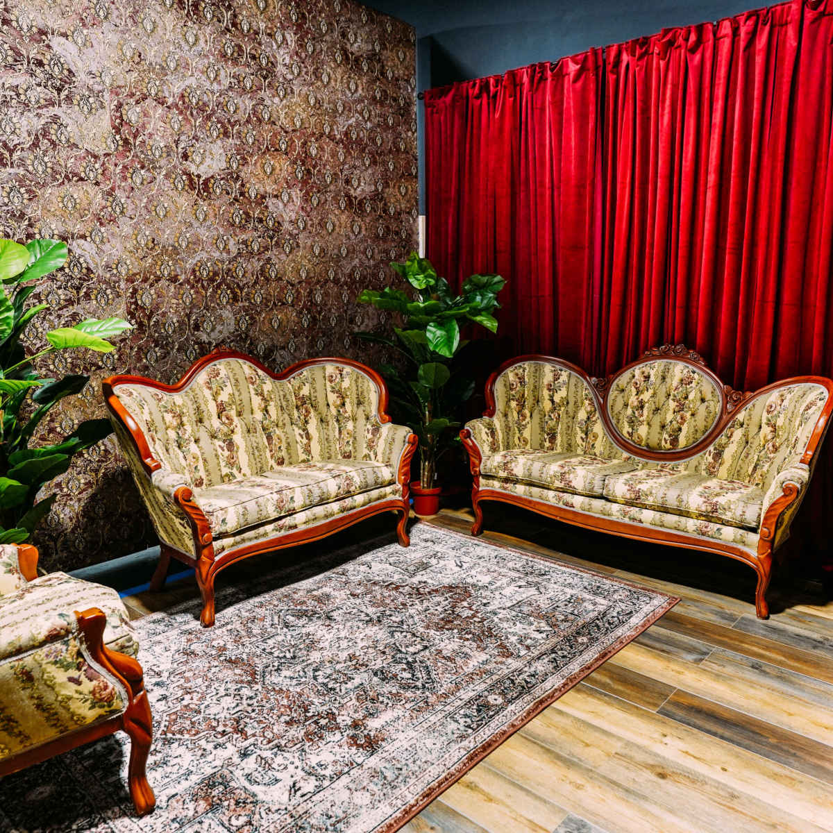 A cozy sitting area with two ornate sofas facing each other, a small side table, a patterned rug, and a backdrop of red curtains and decorative wallpaper.