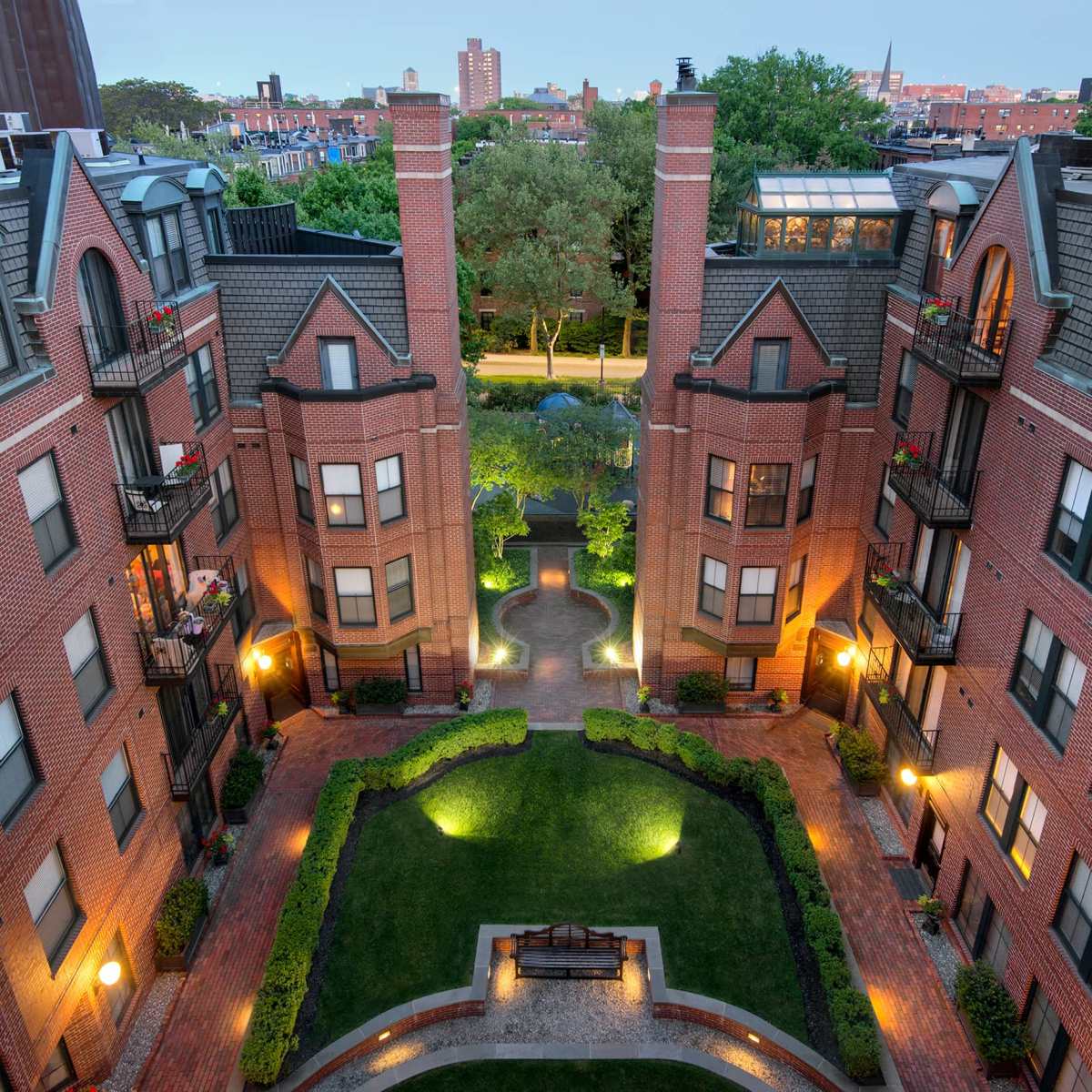 A landscaped courtyard surrounded by brick apartment buildings, with pathways lined by lights and trees, set against a backdrop of city buildings.