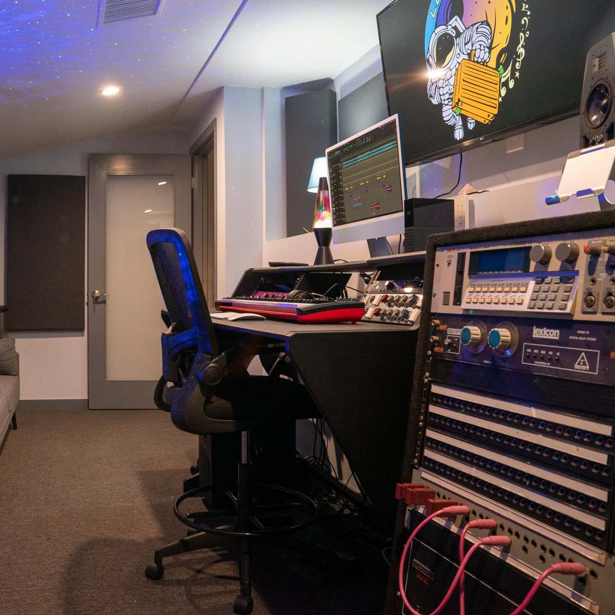 A recording studio with a keyboard, computer monitors, audio equipment, a black office chair, and a gray sofa against the wall.