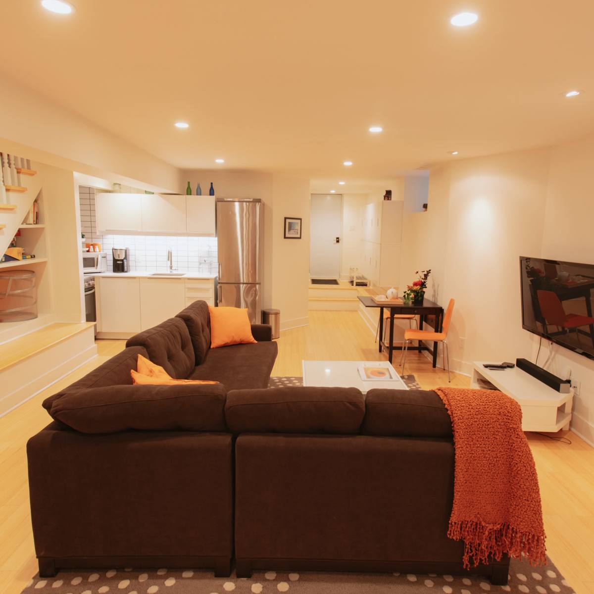 A modern living room features a dark sectional sofa with orange accents, a coffee table, and a television, with a staircase and kitchen visible in the background.