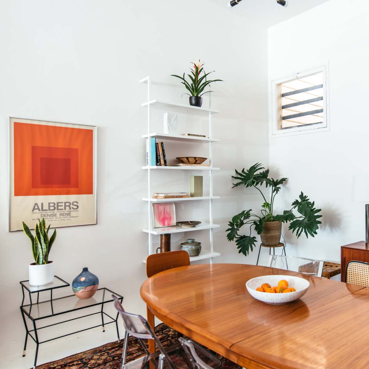 A modern dining area featuring a round wooden table, chairs, and a minimalist shelf with plants and decor against a white wall.