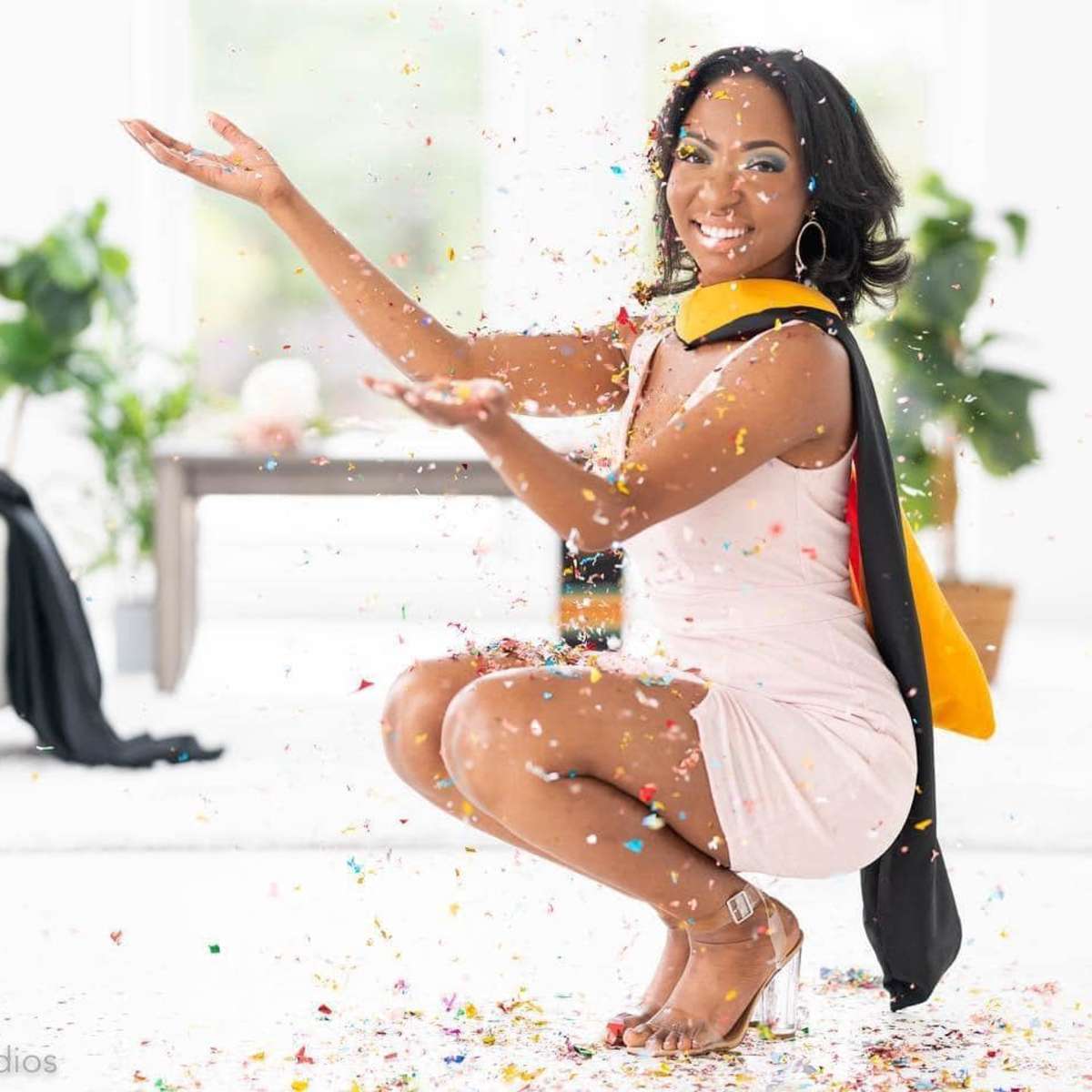 A woman in a light dress and graduation cape crouches down with her arms outstretched, celebrating as colorful confetti falls around her.