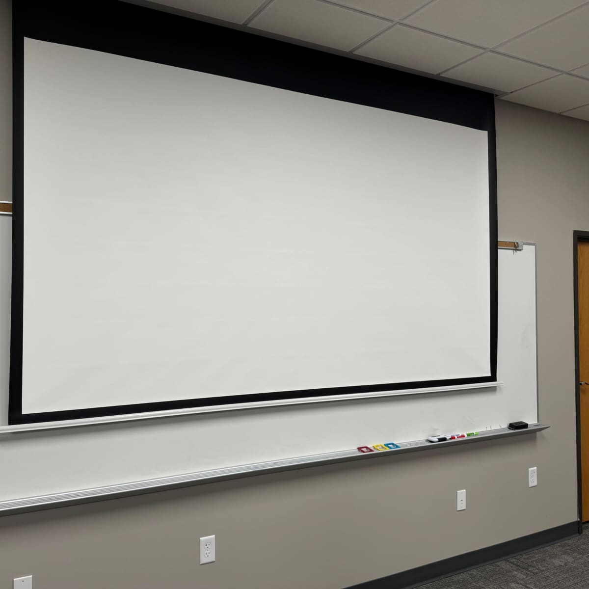 A classroom or presentation room with a blank projection screen mounted on a wall, accompanied by a whiteboard beneath it.