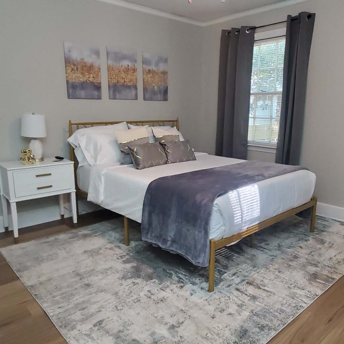 A neatly arranged bedroom featuring a bed with white linens, a gray throw, a nightstand, and three framed artworks on the wall.
