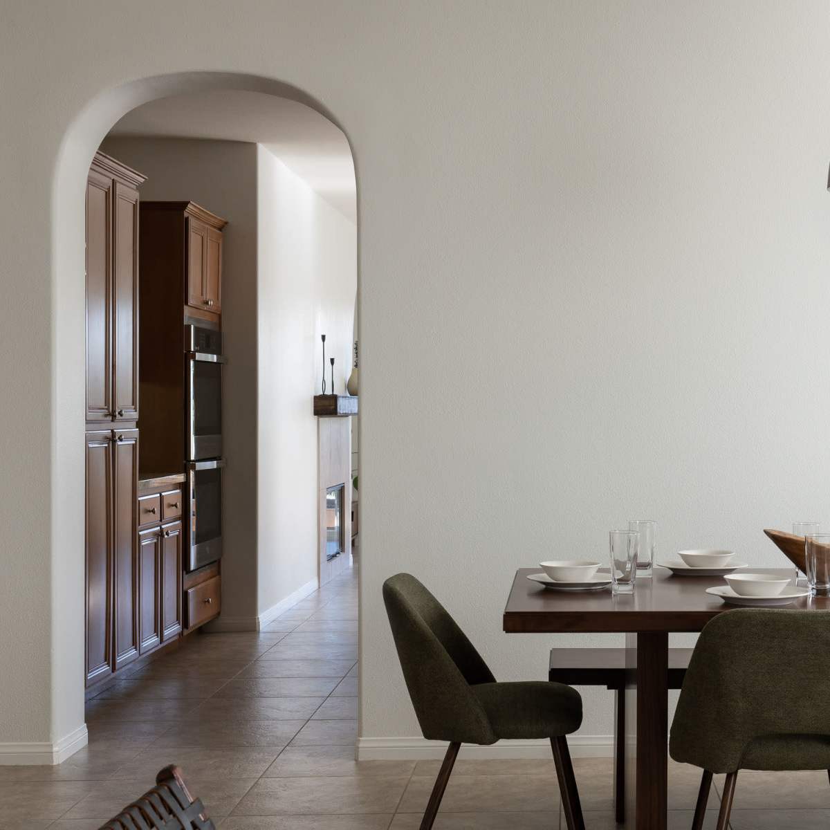 A dining area features a wooden table set with plates and glasses, with an archway leading to a kitchen in the background.