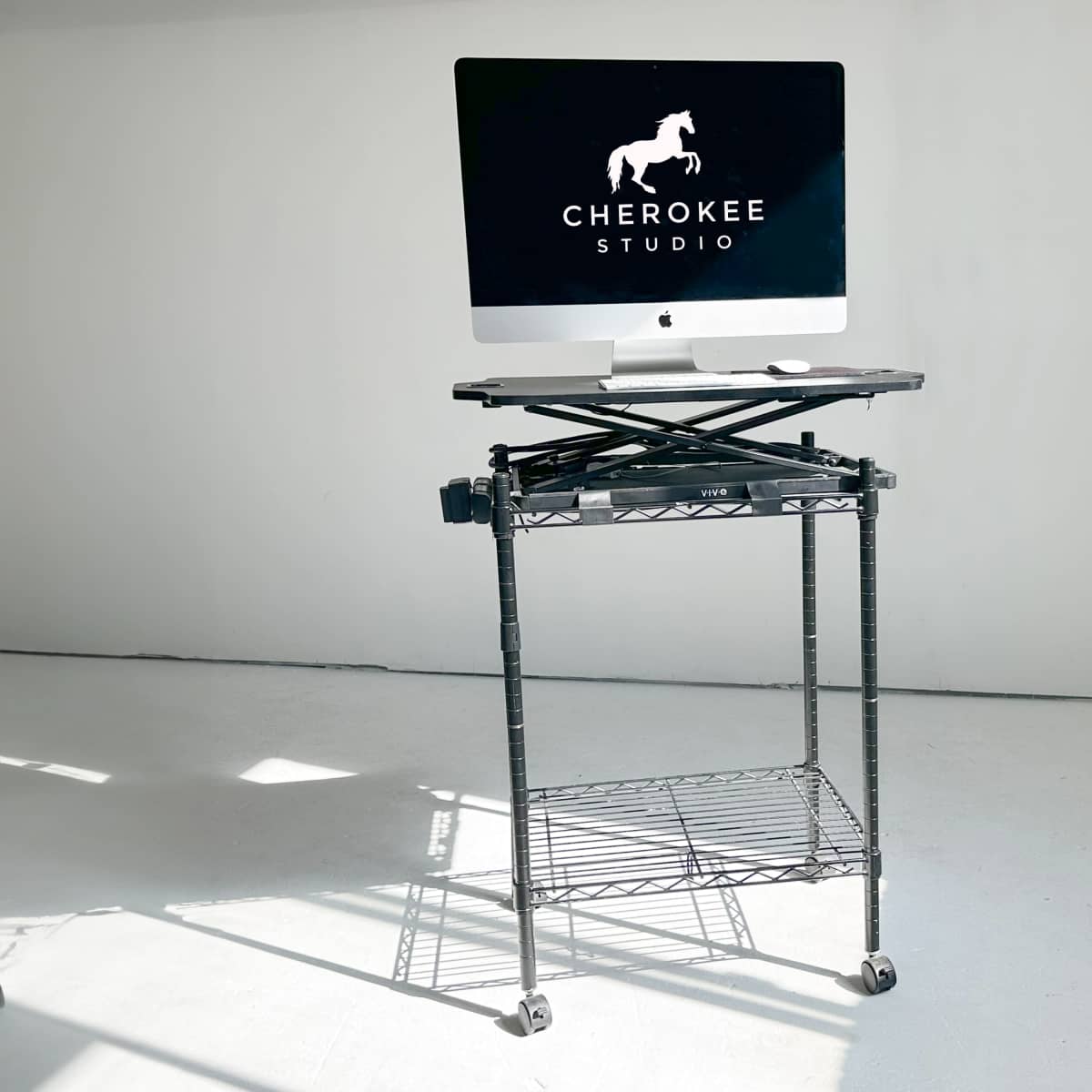 A black mobile cart with a computer monitor and a laptop stand is positioned in a well-lit room with a white backdrop.