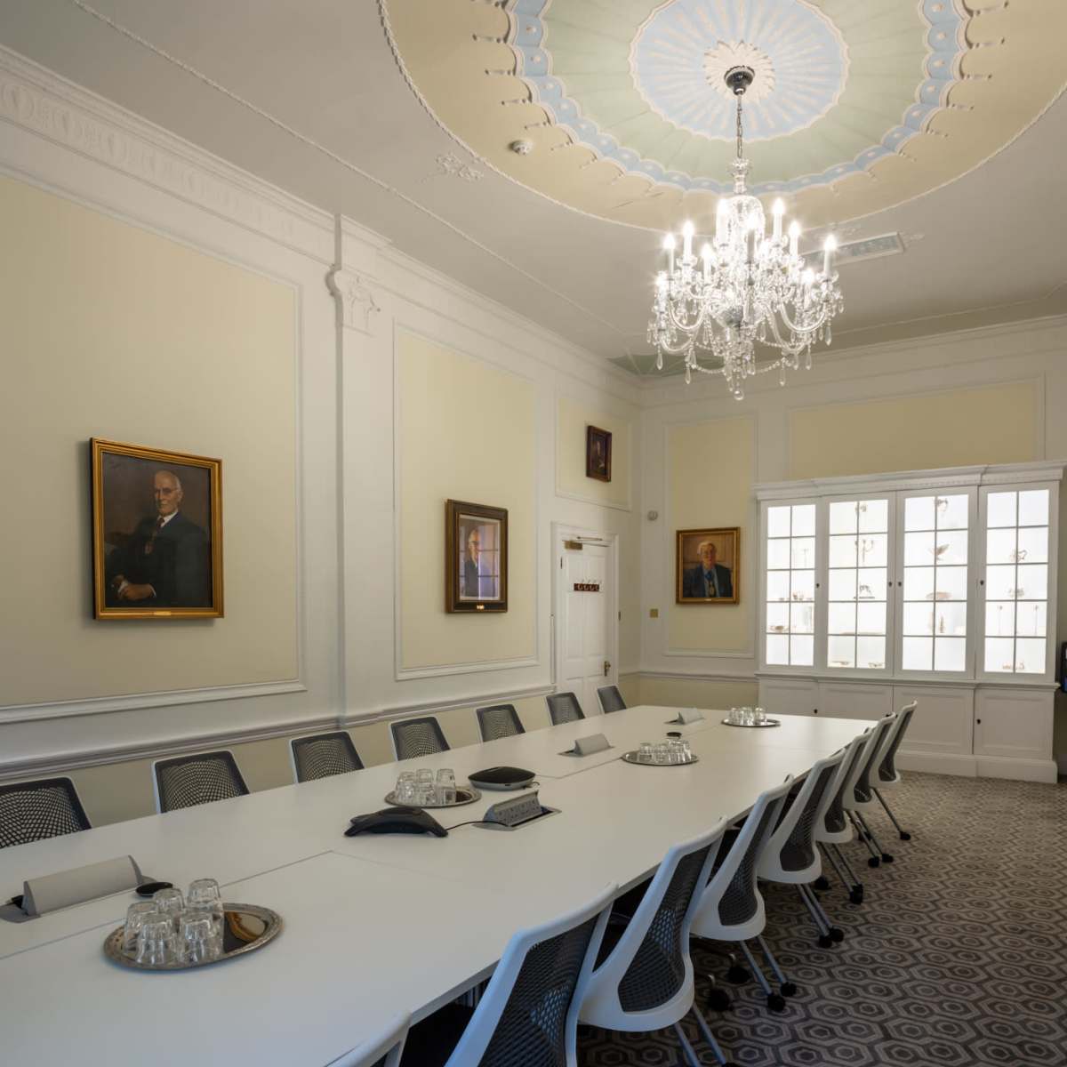 A long, modern conference table is set in a well-lit meeting room adorned with framed portraits and a chandelier.