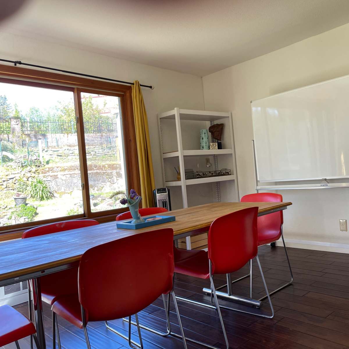 A modern meeting room features a wooden table surrounded by red chairs, with a whiteboard mounted on the wall and a large window overlooking a landscaped area.