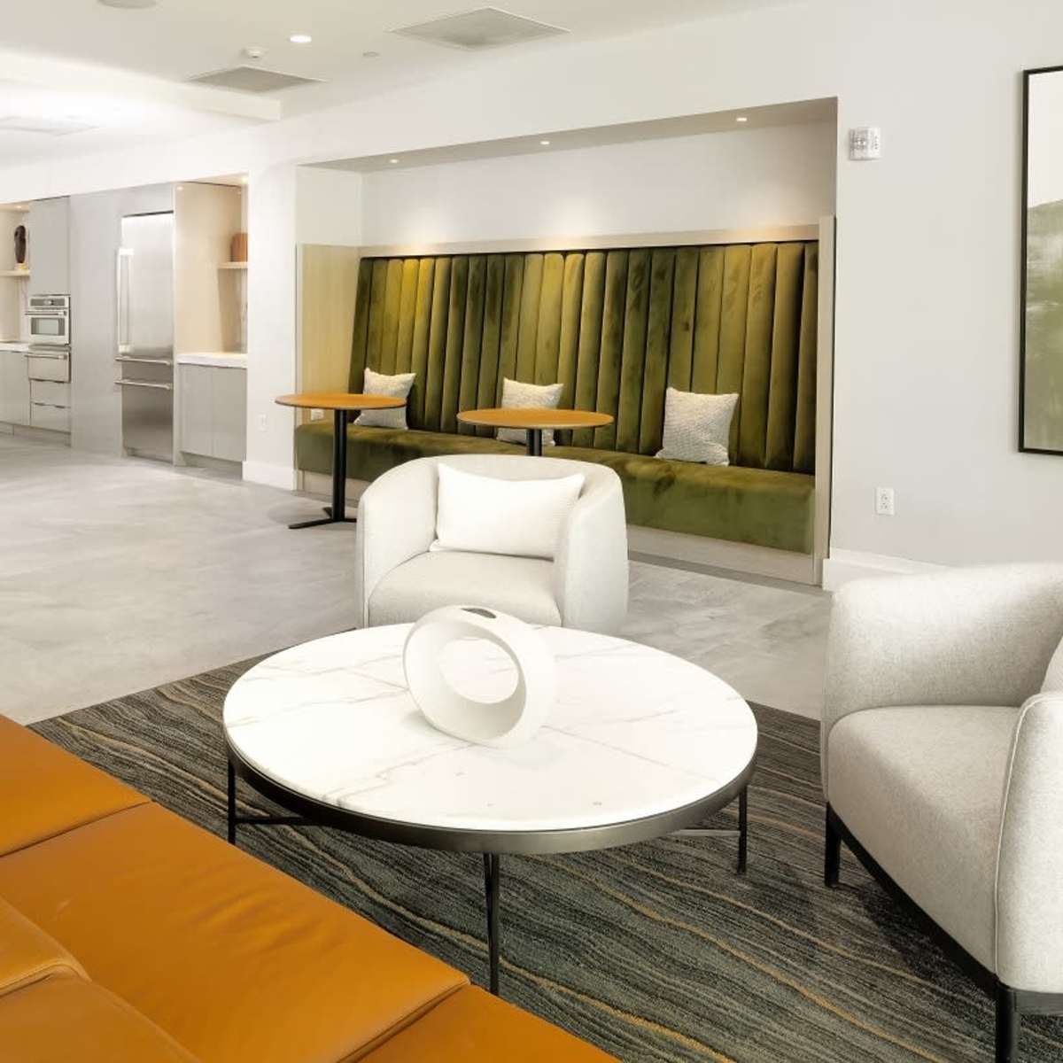 A modern interior space featuring a seating area with a brown leather sofa, a round marble coffee table, and green upholstered booths in the background, along with a kitchen area equipped with stainless steel appliances.